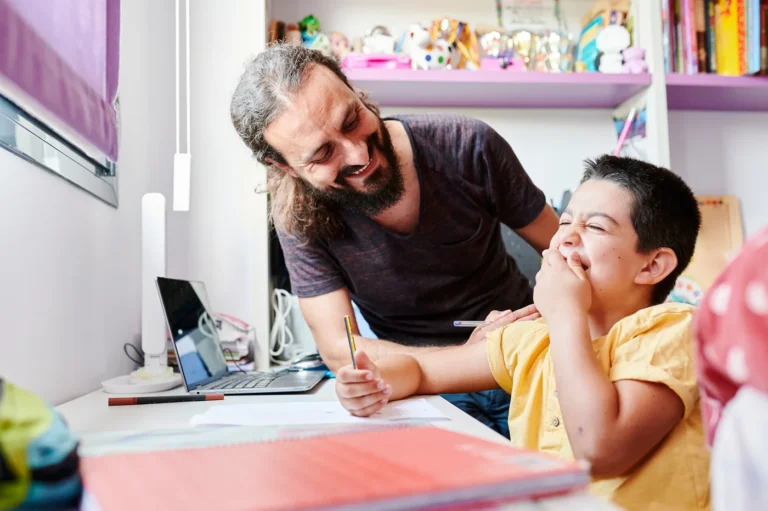 Behavior Analyst smiling and helping a happy young boy write at a desk.